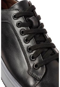 Zapatilla de cuero negra con cordones negros redondos, ojales de metal y una suela de goma transparente. Superficie lisa con un detalle de textura mínimo.