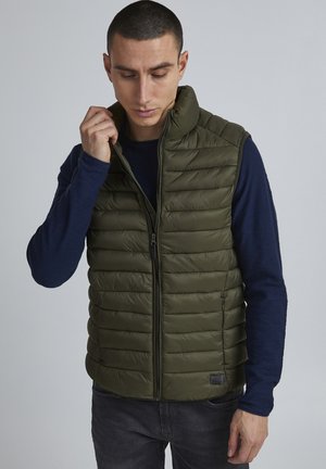 Olive green puffer vest with horizontal stitching, high collar, and front zipper. Worn over a navy long-sleeve shirt, featuring a subtle logo.