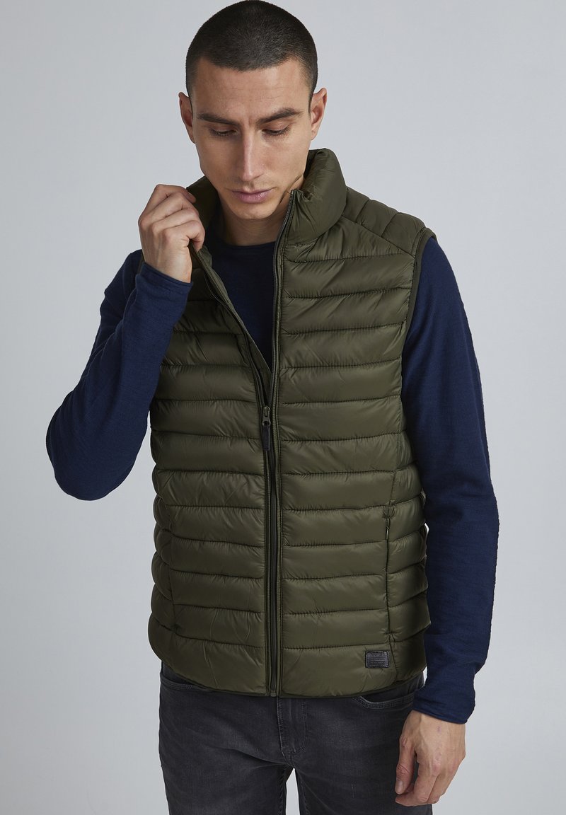 Olijfgroen puffer vest met horizontale stiknaden, hoge kraag en voorkeursrits. Draag over een marineblauw shirt met lange mouwen, met een subtiele logo.