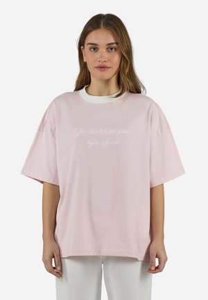 Fiatal nő hosszú barna hajjal, túlméretezett világos rózsaszín pólót visel fehér felirattal: "Nem tudod levenni rólam a szemed" és fehér nadrágot.