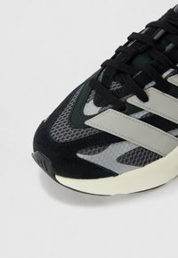 Sneaker avec une tige noire et grise, fabriquée en matériaux en mesh et en daim, avec une semelle intermédiaire blanche épaisse et des rayures grises proéminentes sur les côtés.