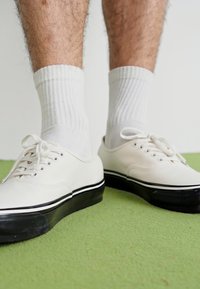 White canvas shoes with black rubber soles, round laces, and a low-top design, worn with white ribbed socks, set on a green surface.