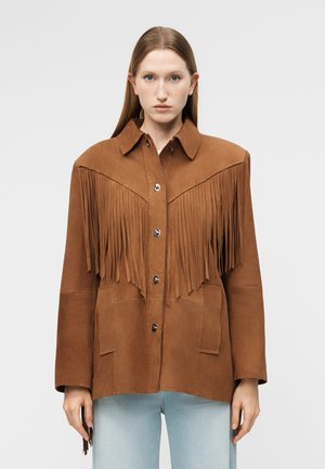 Femme aux cheveux raides châtain clair portant une veste en daim marron avec des détails à franges et un jean bleu clair, face à l'avant sur un fond uni.