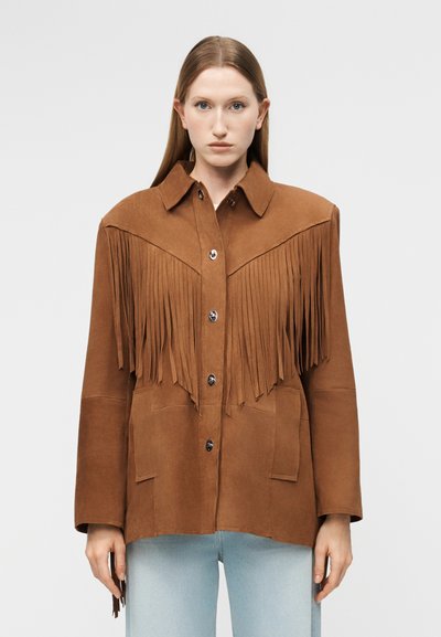 Femme aux cheveux raides châtain clair portant une veste en daim marron avec des détails à franges et un jean bleu clair, face à l'avant sur un fond uni.