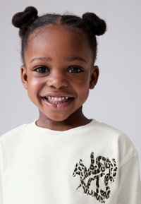 Camiseta de manga larga color crema claro con un diseño texturizado de estampado animal negro en el área del pecho izquierdo. Material de algodón suave, ajuste relajado.