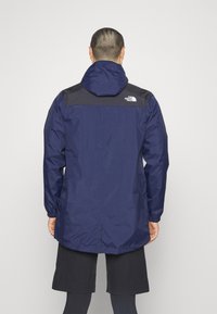 Granatowa wodoodporna kurtka z czarnym panelem na ramieniu, regulowanym kapturem, elastycznymi mankietami i luźnym krojem, z logo na plecach.