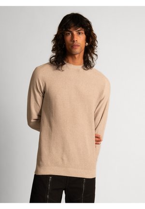 Beige strikket sweater med rund halsudskæring, ribstrikkede manchetter og kant, der har et struktureret mønster og en afslappet pasform. Båret over mørke bukser.