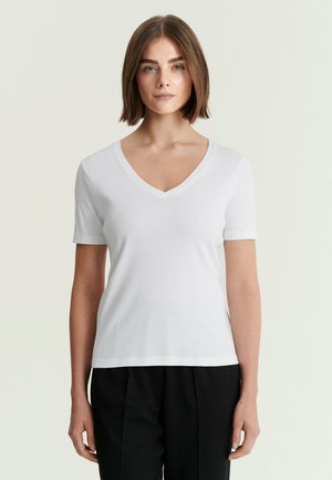 Femme aux cheveux courts châtain portant un t-shirt blanc uni à col en V et manches courtes ainsi qu'un pantalon noir sur un fond clair.