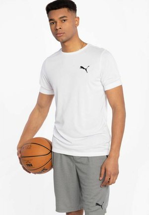 Baltas īsām piedurknēm T-krekls ar melnu Puma logo, apvienots ar pelēkiem sporta šortiem ar melnu logo. Modelis tur basketbola bumbu.