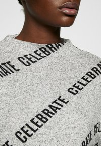 Grå sweatshirt i strukturerad tyg med "CELEBRATE" tryckt i fet svart text diagonalt över designen. Rundad halsringning.