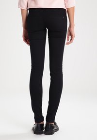 Jeans skinnys negros con una textura suave, cintura de media altura y dos bolsillos traseros, llevados con zapatos negros. La tela parece elástica y ajustada.