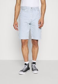 Man som bär vita och ljusblå vertikalt randiga shorts, vita strumpor och svarta högskaftade sneakers, stående mot en enfärgad bakgrund.