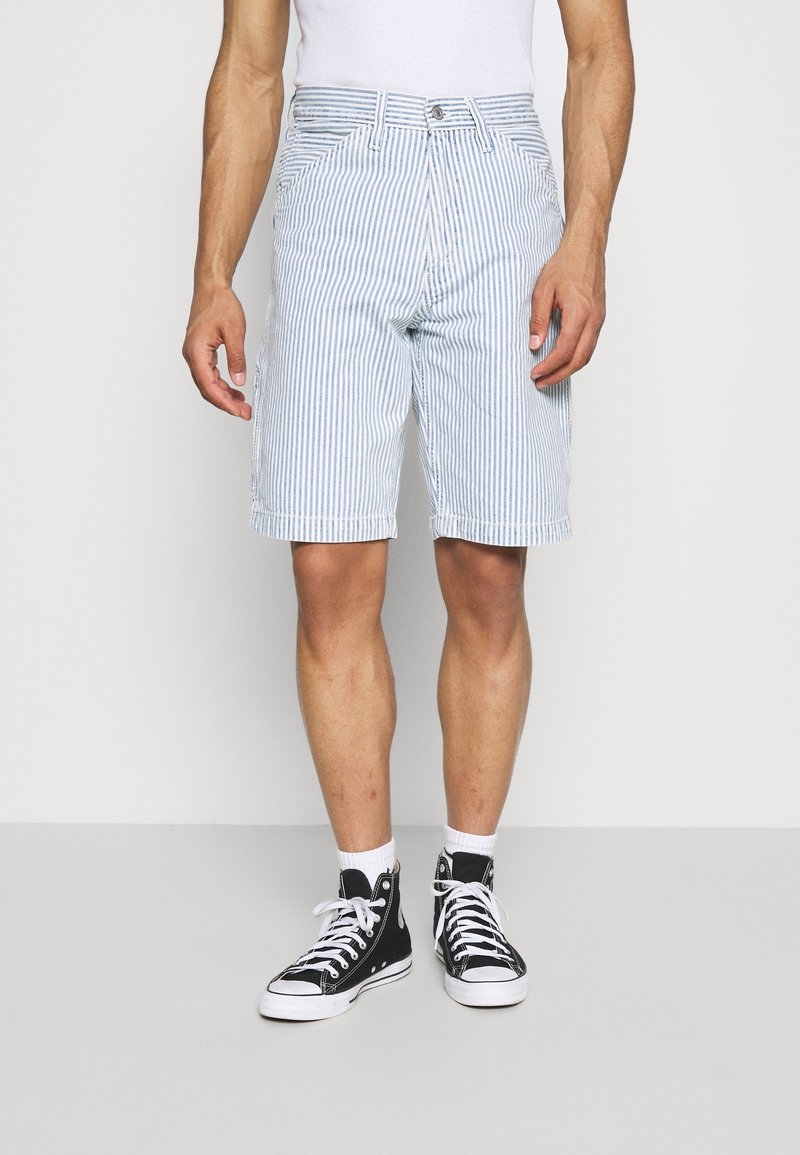 Man som bär vita och ljusblå vertikalt randiga shorts, vita strumpor och svarta högskaftade sneakers, stående mot en enfärgad bakgrund.