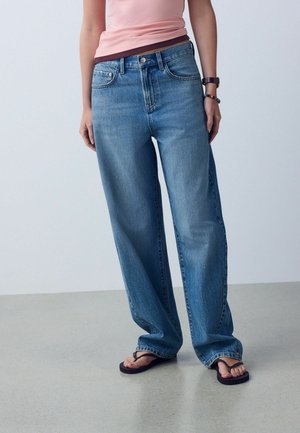 Person iført løse blå jeans, en lyserød ærmeløs top, et ur og armbånd på højre håndled og sorte flip-flops.