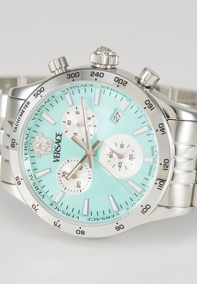 Silverfärgad klocka i rostfritt stål med en turkos urtavla, tre subdials och en lejonsymbol. Utrustad med en tachymeter och texturerade timmarkörer.