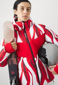 Veste de ski rouge et blanche rembourrée avec des motifs en vagues, dotée d'un col haut et d'une fermeture éclair noire. Un casque de ski gris et un bâton de ski sont tenus.