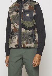 Chaleco de forro polar en patrón de camuflaje con mangas negras, cierre frontal y bolsillo; superficie texturizada con tonos verdes, marrones y beiges.