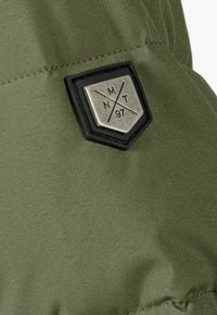 Tejido verde con una textura suave presenta un parche en forma de escudo negro con un diseño en relieve plateado y las letras "MNT" y "97."