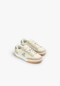 Beige Mesh-Sneaker mit grauen Wildlederelementen, weißen Schnürsenkeln, einer blauen Fersenkappe und einem Totenkopf-Logo an der Seite. Orange Laufsohle.