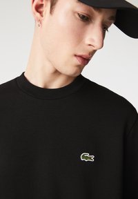 Joven vestido con un sudadera negra de cuello redondo con un logo de cocodrilo verde y una gorra negra, mirando hacia abajo de lado.