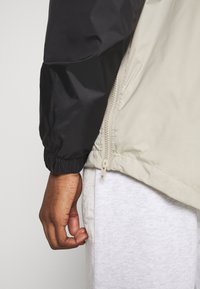 Giacca bicolore con manica nera e corpo beige, con polsini elastici, zip laterale e una texture di tessuto liscia e resistente all'acqua.