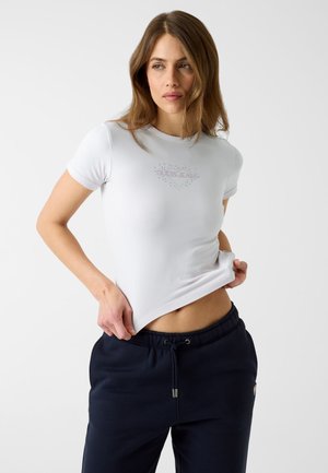 Woman with long brown hair wearing a white fitted t-shirt and navy blue drawstring pants, lifting the shirt slightly with one hand.