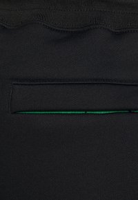 Schwarzer Stoff mit glatter Textur, der über eine schmale, versteckte Tasche mit grünem Akzent am Rand verfügt. Der obere Rand hat ein gewelltes Design.