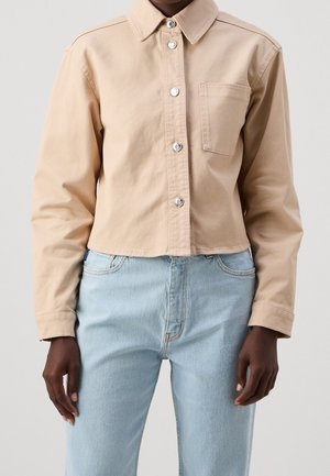 Camisa beige recortada con botones y bolsillo en el pecho, usada con vaqueros de tiro alto en azul claro en una persona contra un fondo liso.