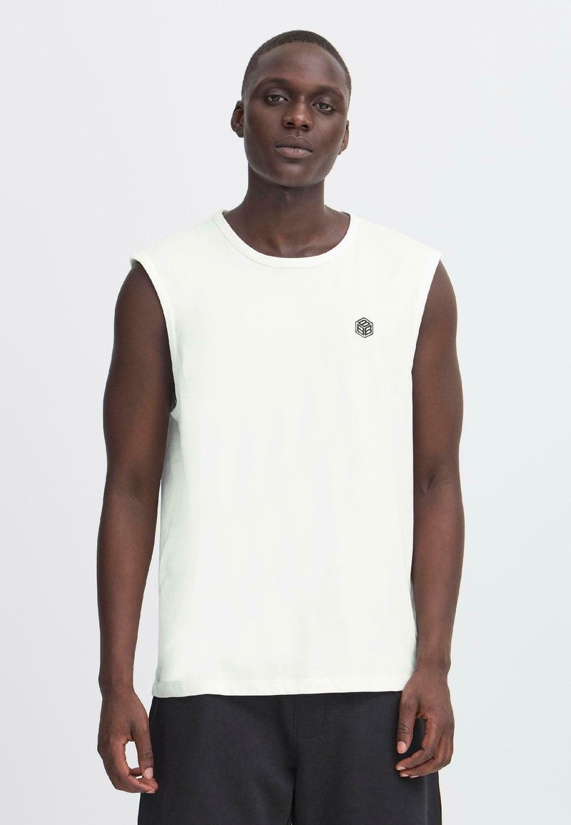 Canotta bianca in cotone senza maniche con collo tondo e piccolo logo geometrico nero sul lato sinistro del petto, abbinata a pantaloni corti scuri.