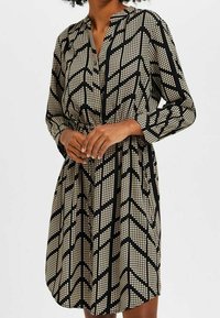 Femme portant une robe noire à manches longues avec des motifs chevron à pois blancs géométriques, taille cintrée, se tenant contre un fond clair uniforme.