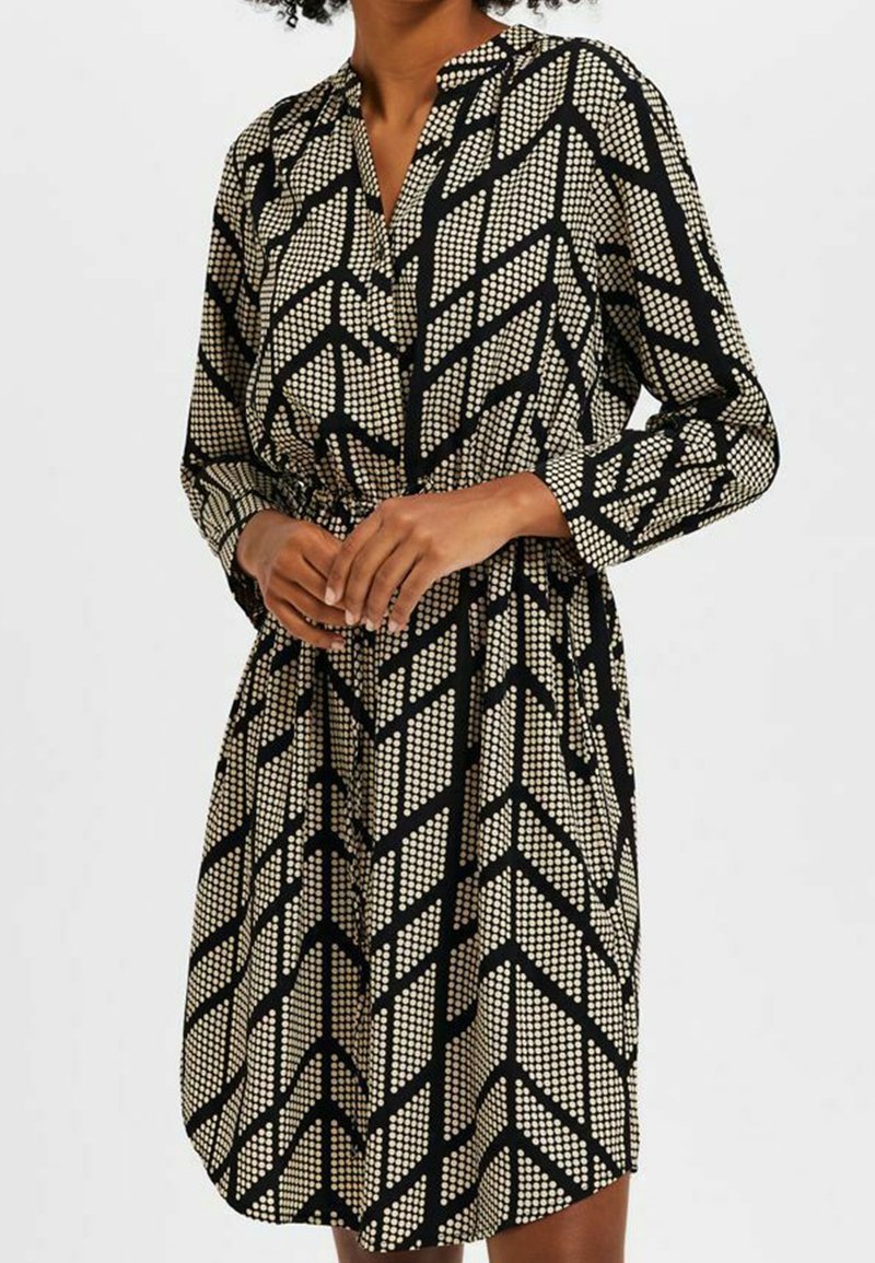 Femme portant une robe noire à manches longues avec des motifs chevron à pois blancs géométriques, taille cintrée, se tenant contre un fond clair uniforme.