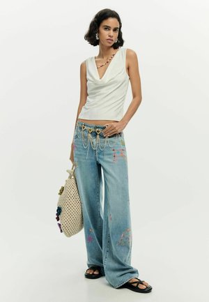 Donna con top bianco senza maniche, jeans denim a gamba larga ricamati con cintura a catena, che tiene una borsa all'uncinetto e indossa sandali neri su sfondo bianco.