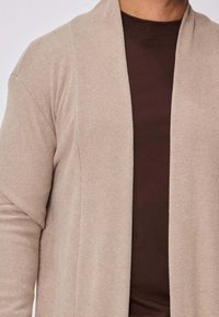 Cardigan en tricot beige à devant ouvert et manches longues, fabriqué en matière douce et texturée, superposé à une chemise marron.