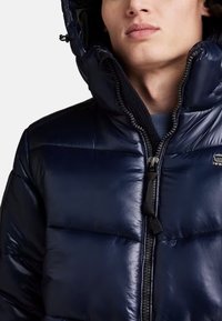 Modra puffer jakna z bleščečo površino, zadrgo, kapuco in strukturirano šivanje, z logotipom na predelu prsnega koša.