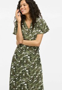 Zavinovací šaty v zelené barvě s motivem listů. Krátké rukávy, výstřih do V a detaily s vázáním v pase. Lehčená látka s jemnou texturou.