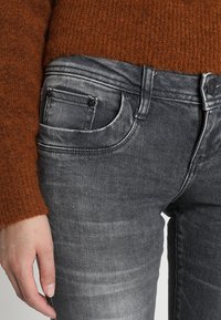Jeans de mezclilla gris con un diseño ajustado, que presentan un estilo de cinco bolsillos y costuras visibles. Combinados con un suéter marrón texturizado.
