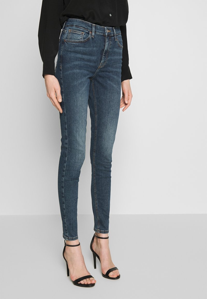 Jean en denim foncé skinny avec une taille mi-haute, comportant cinq poches et une légère décoloration. Associé à des sandales à talons hauts noires à lanières.