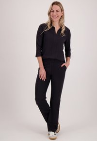 Glimlachende vrouw met blond haar staat met één hand in haar zak, gekleed in een zwart shirt met lange mouwen, zwarte broek en witte sneakers.