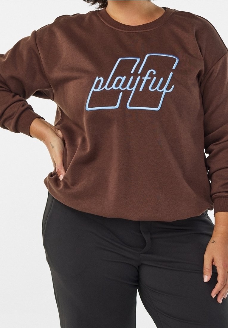 Personne portant un sweat-shirt marron avec un texte contour bleu "playful" et un pantalon noir, une main sur la hanche et l'autre le long du corps.