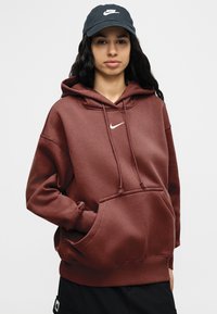 Νεαρός με μακριά σκούρα μαλλιά που φοράει μπορντό φούτερ Nike και μαύρο καπέλο Nike, στεκόμενος με το ένα χέρι στην τσέπη του φούτερ.