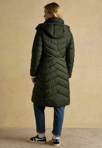 Olivovo zelená páperová bunda s chevronovým prešívaním, kapucňou a zipsovým uzáverom. Má priliehavý pás a bočné vrecká, nosená cez modré džínsy.