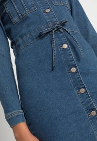 Farkku mekko, jossa on istuva vyötärö, pitkät hihat ja nappisuljenta. Ominaisuuksiin kuuluu kaksi rintataskua ja solmittava vyö. Väri: tummansininen.