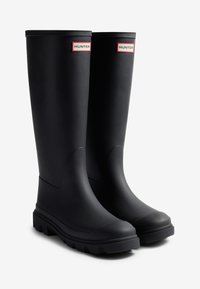 Bottes de pluie en caoutchouc noir avec une finition mate lisse, dotées d'un petit talon et d'étiquettes de marque décoratives rouges et blanches en haut.