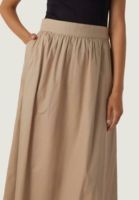 Ljusbeige maxi kjol med en platt midjeband, rynkad textur och sidofickor. Tillverkad av ett mjukt tyg med en slät yta.