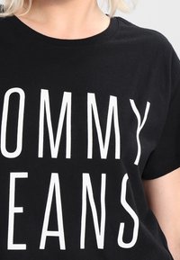 Osoba oblečená v černém tričku s velkými bílými písmeny, která na hrudi říkají "TOMMY JEANS".