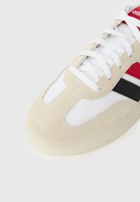 Beige ruskinds sneakers med hvide mesh-detaljer, røde og sorte striber samt hvide snørebånd. Rund tå og struktureret gummisål.
