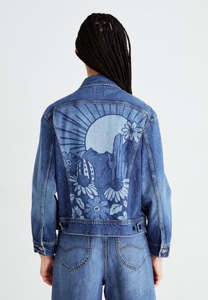 Osoba z długimi warkoczami ubrana w niebieską dżinsową kurtkę i dżinsy, na plecach kurtki grafika przedstawiająca słońce, góry, kaktusy i kwiat.