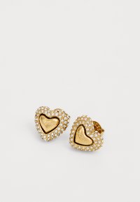 Guld hjärtformade örhängen utsmyckade med rhinestones, med en slät mittdel där det står "GUESS!" i mitten, vilket ger ett texturerat utseende.