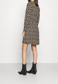 Robe noire à fleurs avec manches longues, présentant un détail froncé au dos. Portée avec des bottines noires, mettant en valeur une texture lisse et une coupe ajustée.