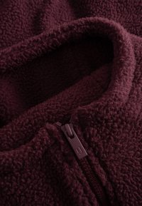 Burgundin värinen fleece-takki, jossa on pehmeä, teksturoitu kangas, vetoketjukiinnitys ja rullakaulus. Huomattava vetoketjun väri vastaa kangasta.